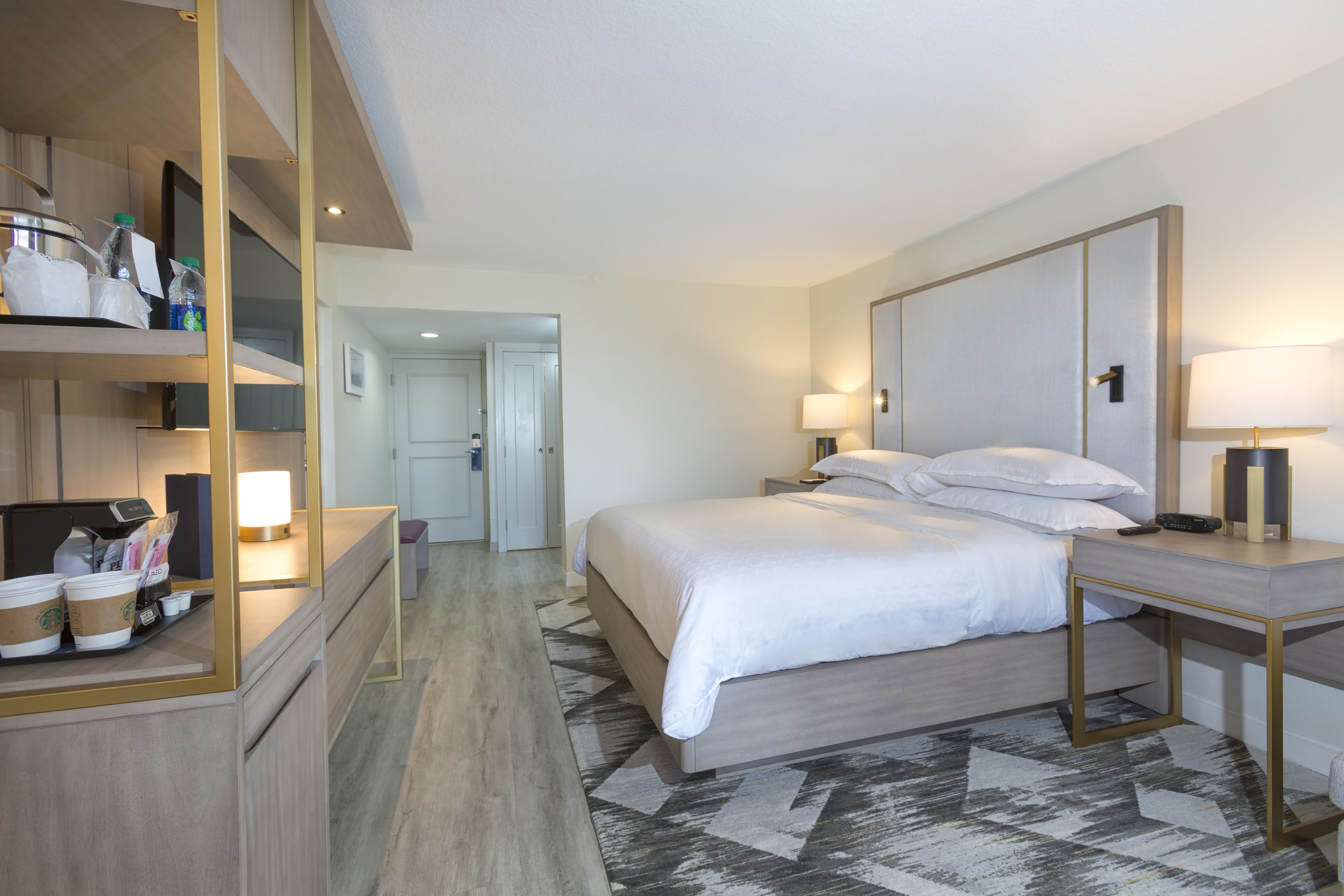 A hotel room with a bed, bedside tables, a desk with a lamp, a mirror, and a coffee station.