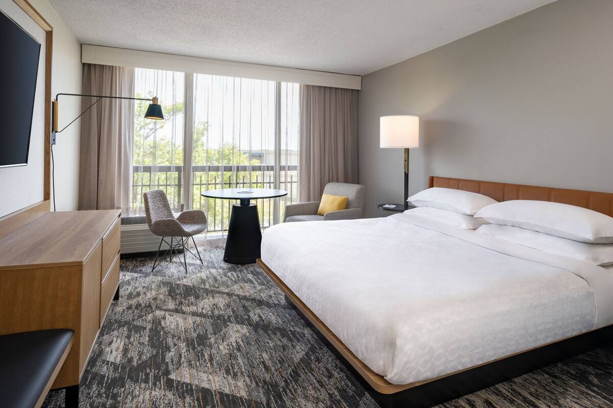 A hotel room with a large bed, a desk, a chair, a small table, and a window with curtains.
