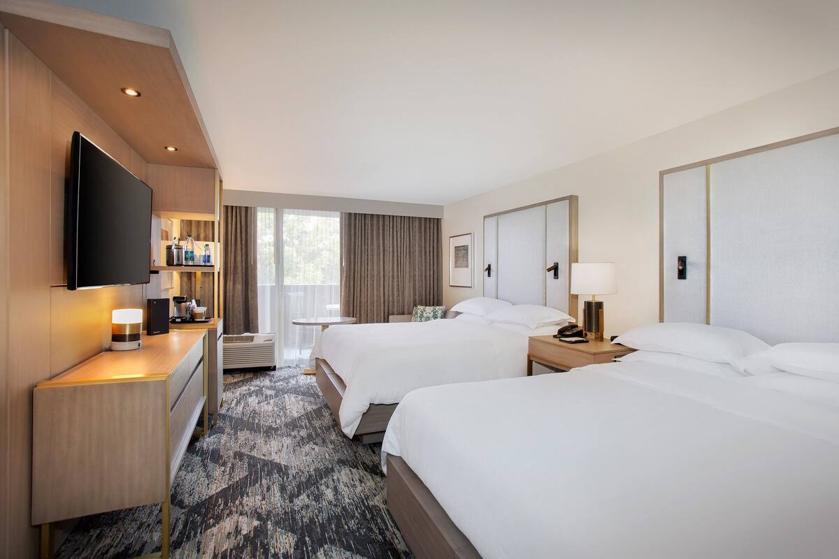 A hotel room with two beds, a desk with a flat-screen TV, a minibar, and a large window with curtains.