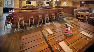 Una zona de bar con taburetes de madera, una barra y un área de comedor con mesas preparadas para dos.