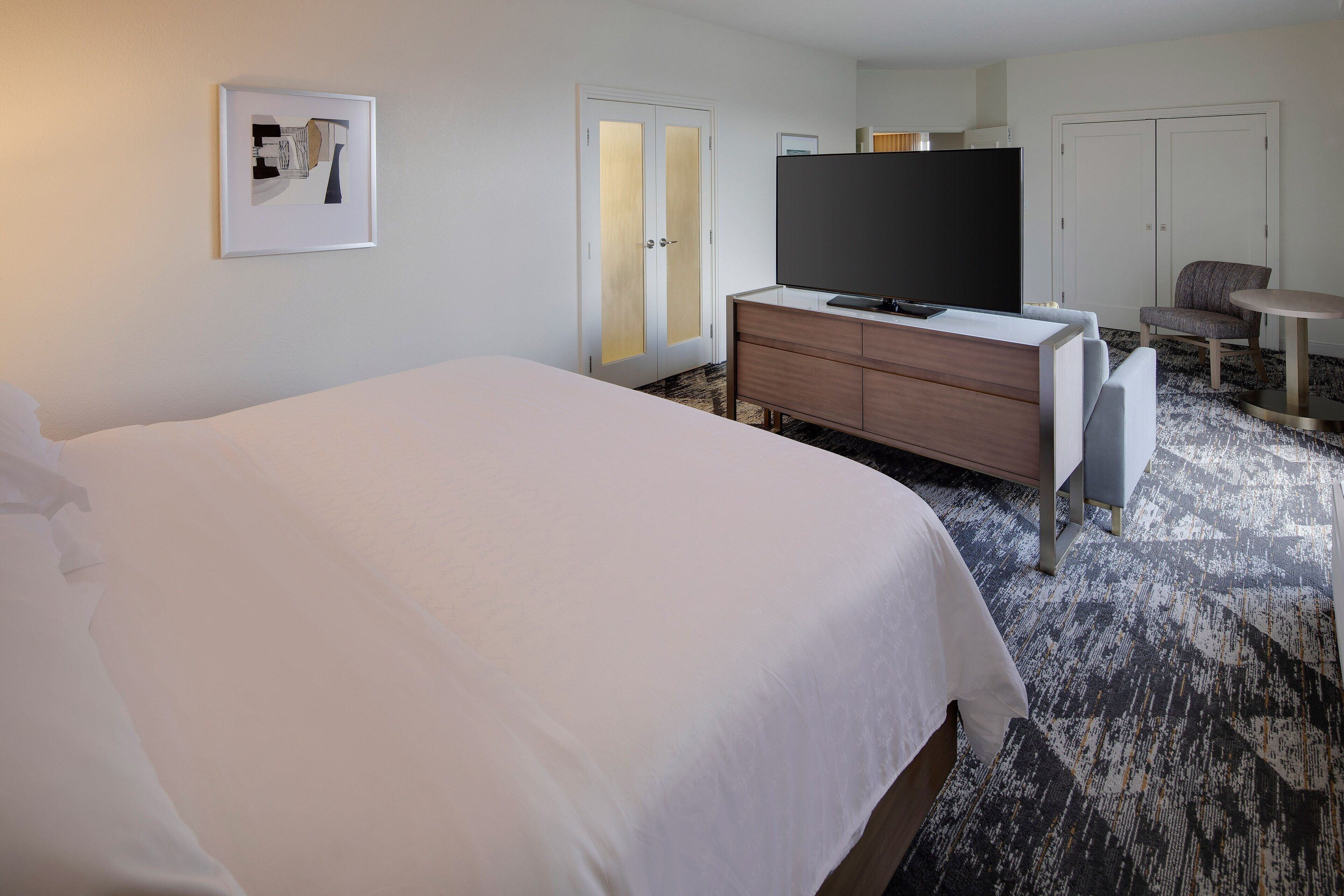 A hotel room with a large bed, a flat-screen TV, a wooden dresser, a gray armchair, and a small table.