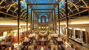A spacious indoor dining area with a high ceiling and a glass structure. Tables are set with candles and tableware.