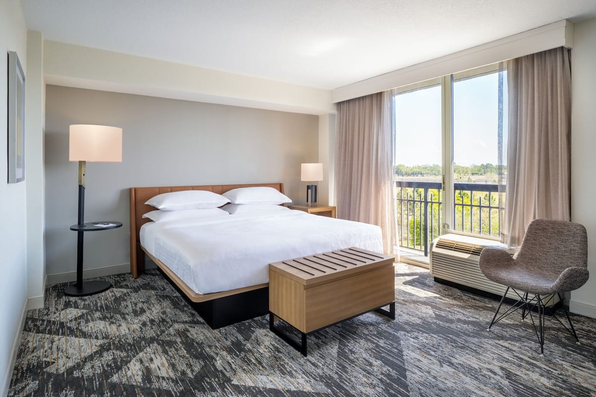A hotel room with a large bed, a bedside table, a chair, a standing lamp, and a view of the outdoors.