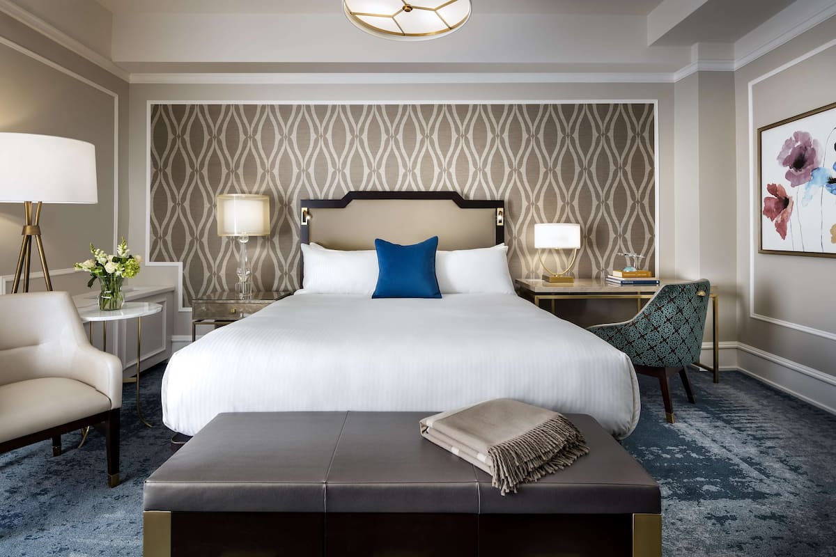 A hotel room with a large bed, two bedside tables, a chair, a bench, and a patterned wallpaper.