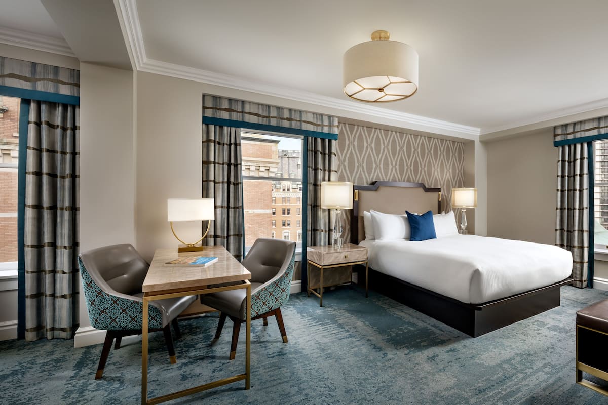 A hotel room with a large bed, two bedside tables with lamps, a desk with two chairs, and a view of the city through the windows.