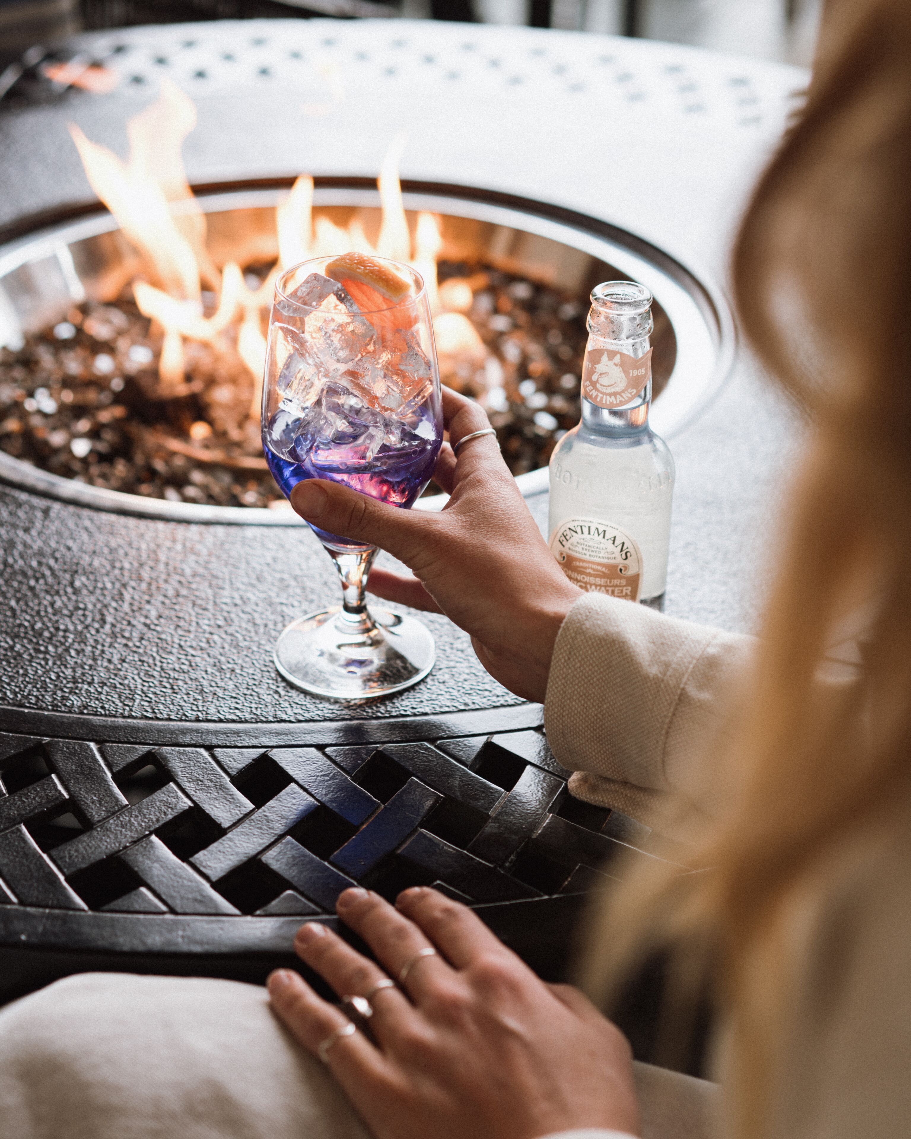 A person holding a glass with a drink and a bottle of liquor on a table with a fire pit in the background.