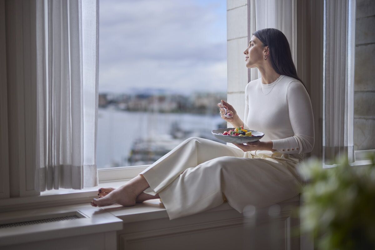 A person sitting by a window with a plate of food, overlooking a body of water.