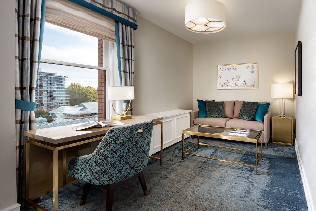 A modern hotel room with a desk, chair, sofa, and a view of the cityscape through large windows.