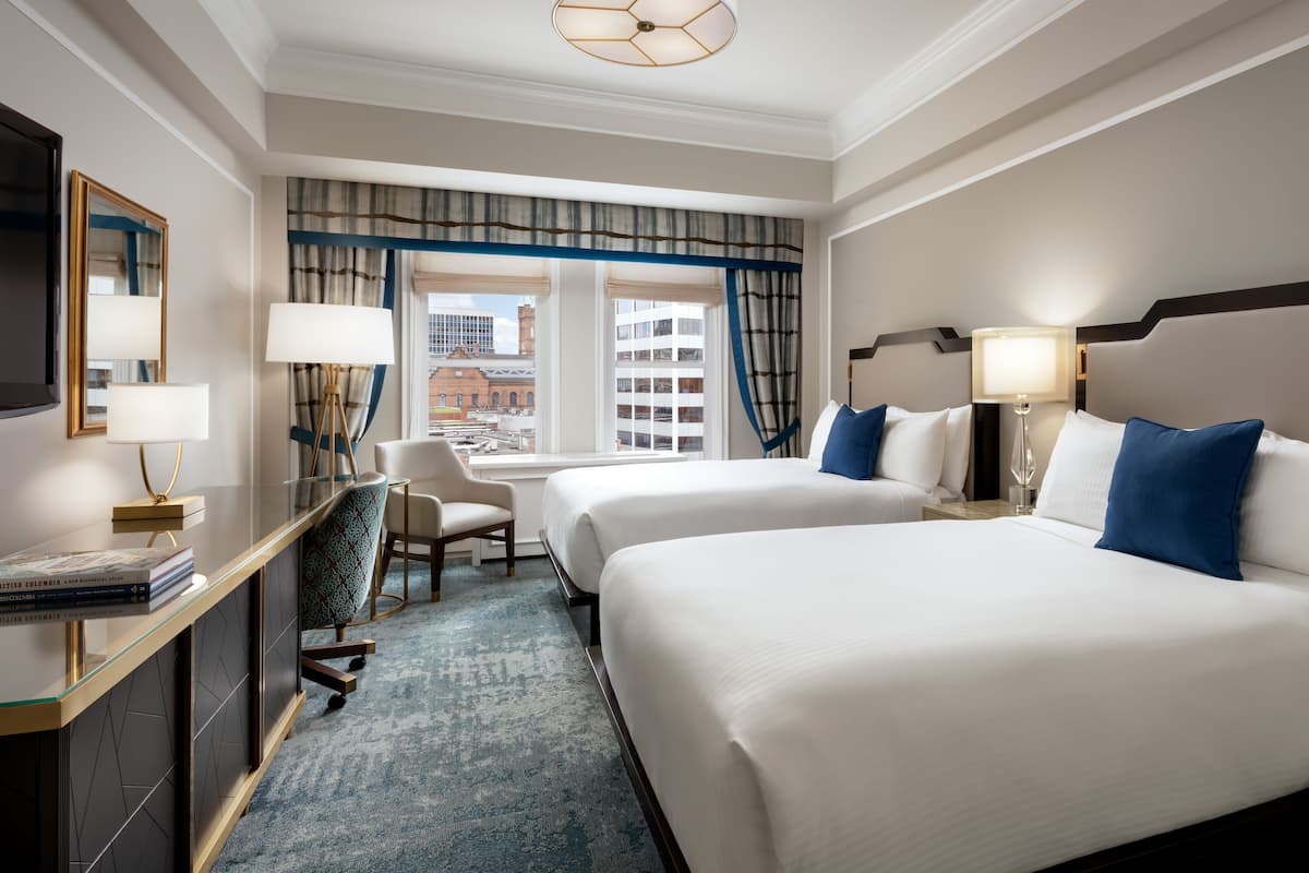 A hotel room with two beds, a desk, a chair, and a view of the city.