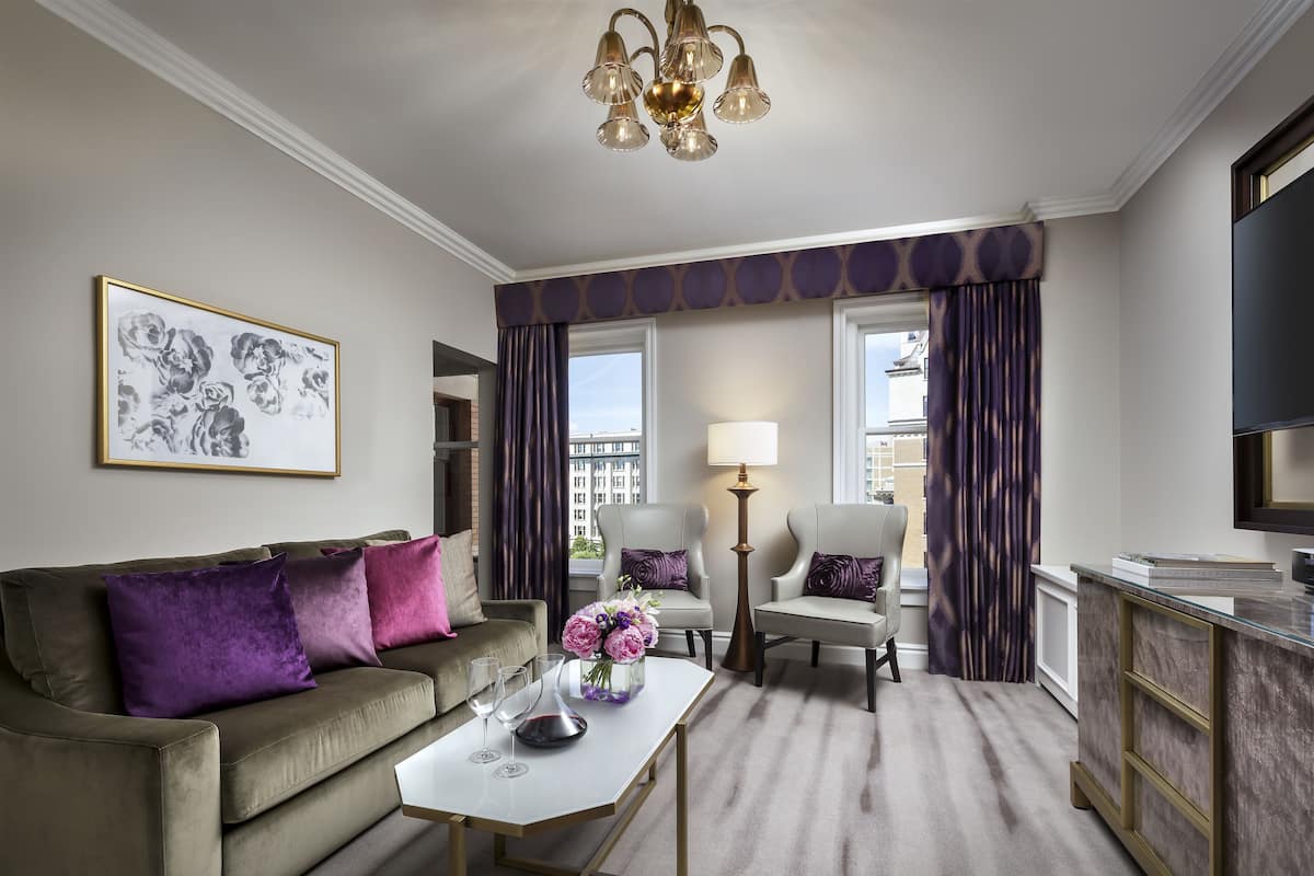 A modern hotel room with a sofa, armchairs, a coffee table, a lamp, and a TV.