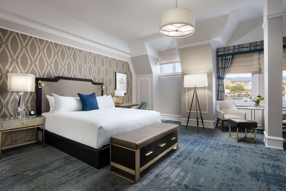A hotel room with a large bed, bedside lamps, a bench, a chair, and a view of the city.
