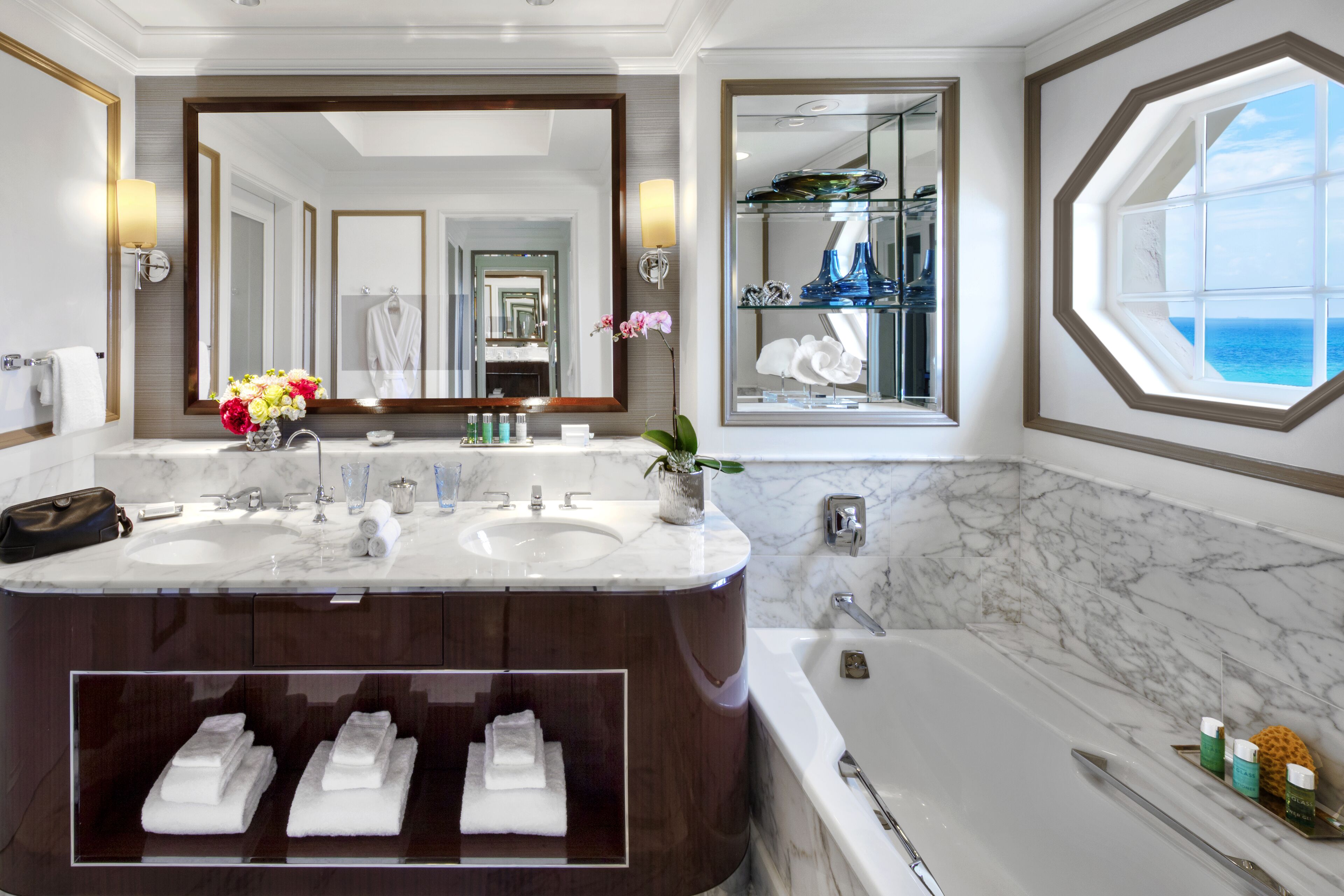 A luxurious bathroom with a large mirror, two sinks, a bathtub, and a view of the ocean.