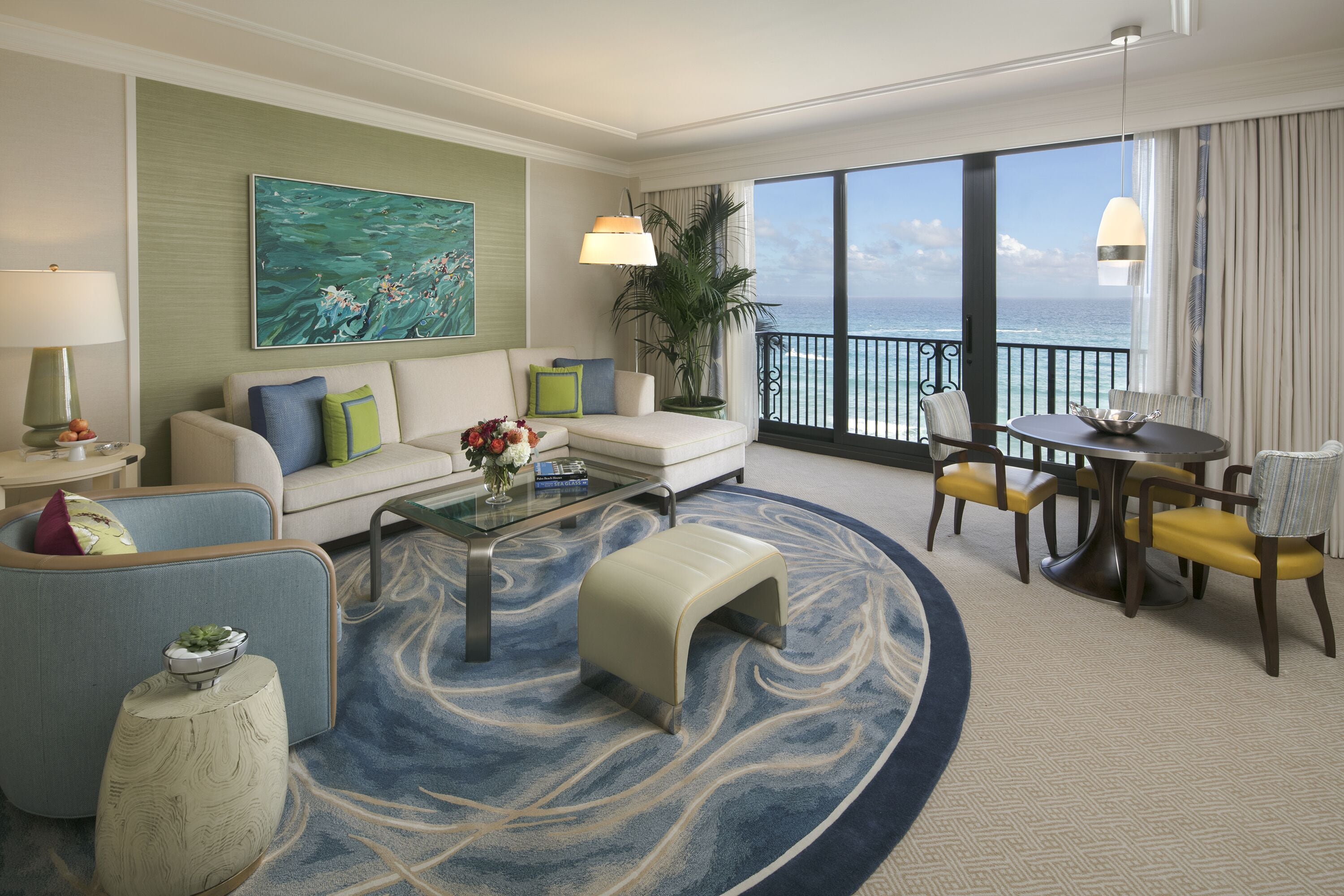 A hotel room with a sofa, armchairs, a coffee table, a dining table with chairs, a view of the ocean, and a large painting on the wall.