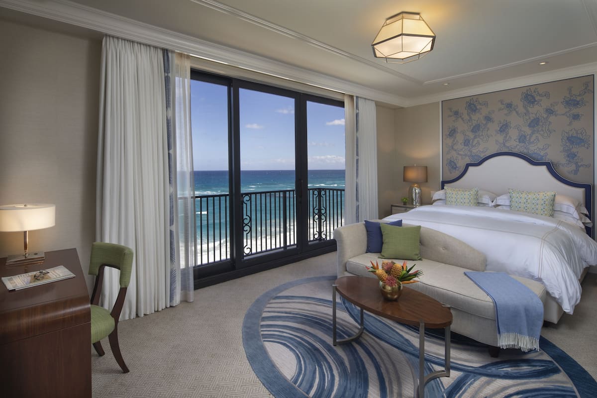 Habitación de hotel con un balcón grande, una cama con ropa de cama blanca, un sofá con una manta azul y un escritorio con lámpara.