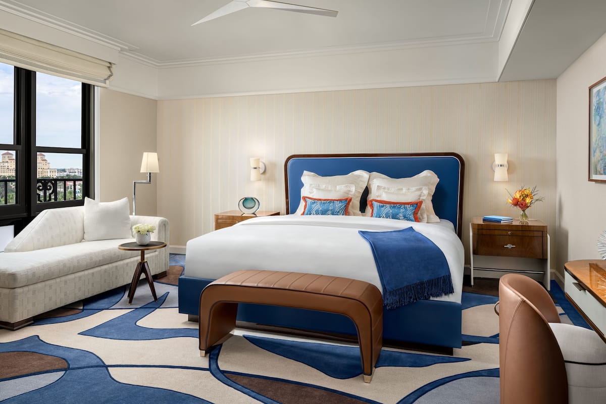 Una habitación de hotel moderna con una cama grande, un sofá, una banca, un escritorio y vistas a la ciudad.