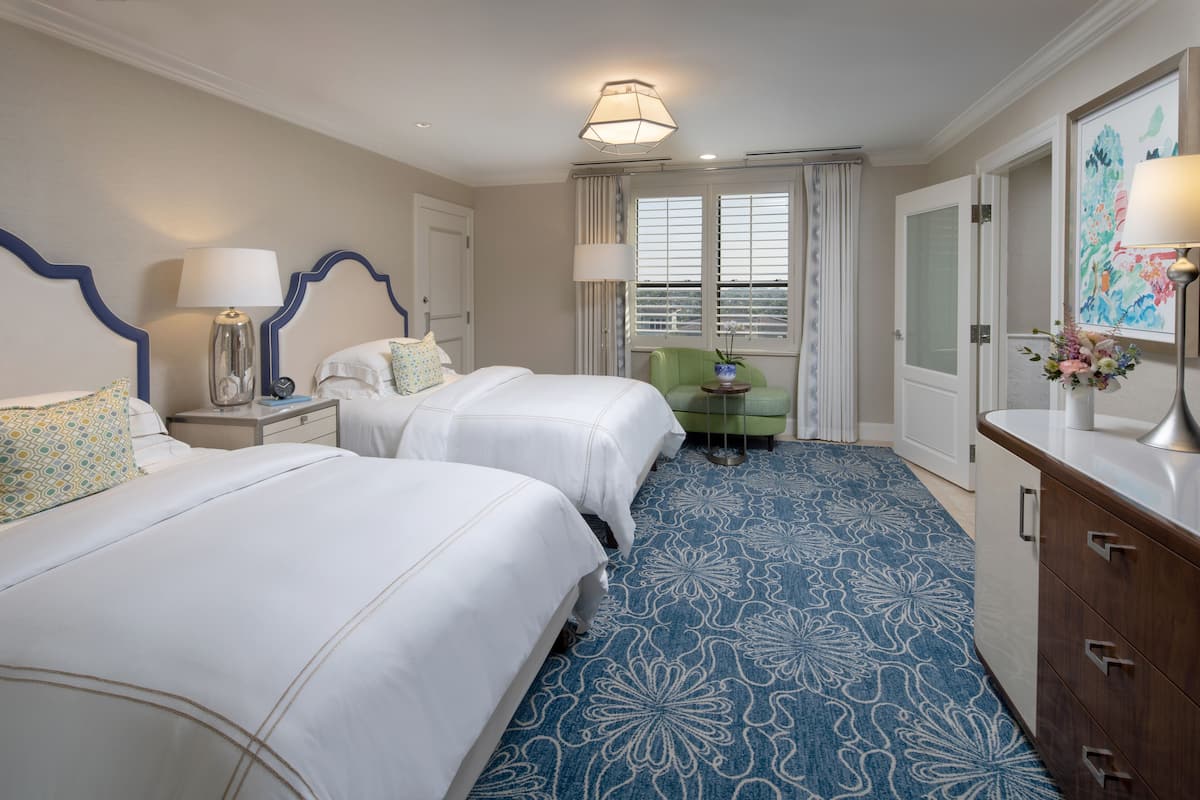 Habitación de hotel con dos camas, una mesita de noche con lámpara, una silla verde, un aparador y una ventana con cortinas.