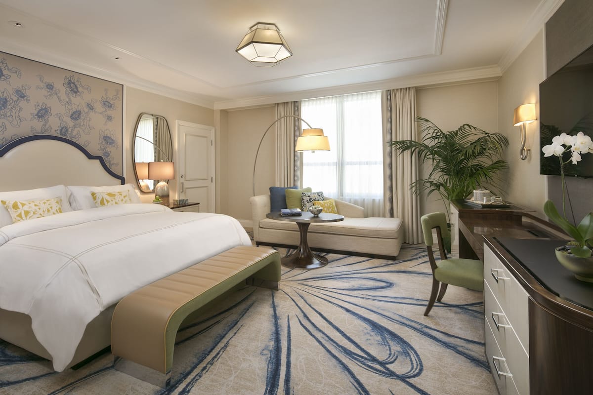 Habitación de hotel con una cama grande, un sofá, un escritorio y una silla. Cuenta con una ventana con cortinas, una lámpara y una planta.