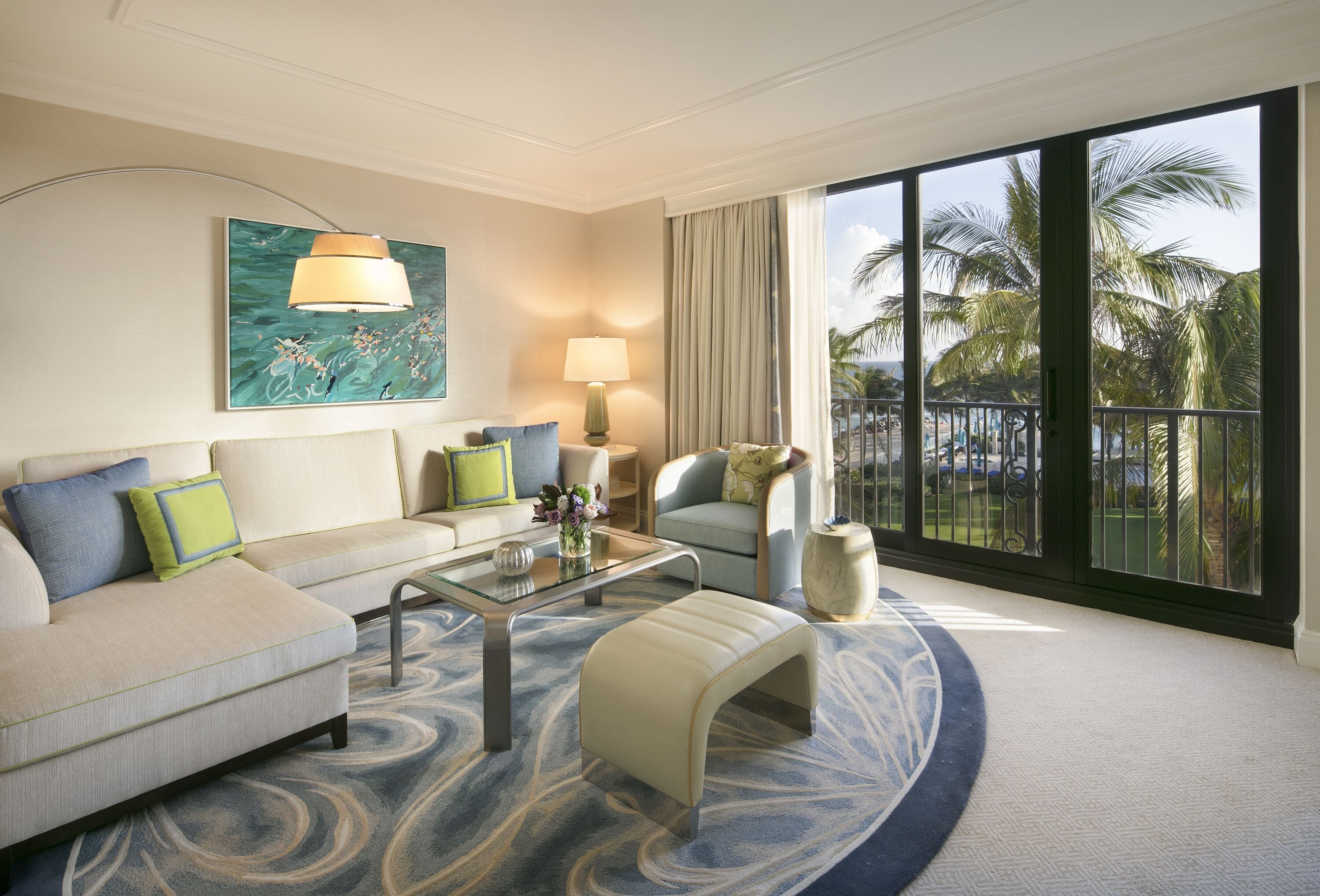 A modern hotel room with a sofa, armchair, and ottoman, a glass coffee table, a lamp, and a large window with a view of palm trees.