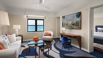 A hotel room with a bed, a desk, a chair, a sofa, a lamp, a vase with flowers, a painting, a window with a view of the sea, and a ceiling fan.