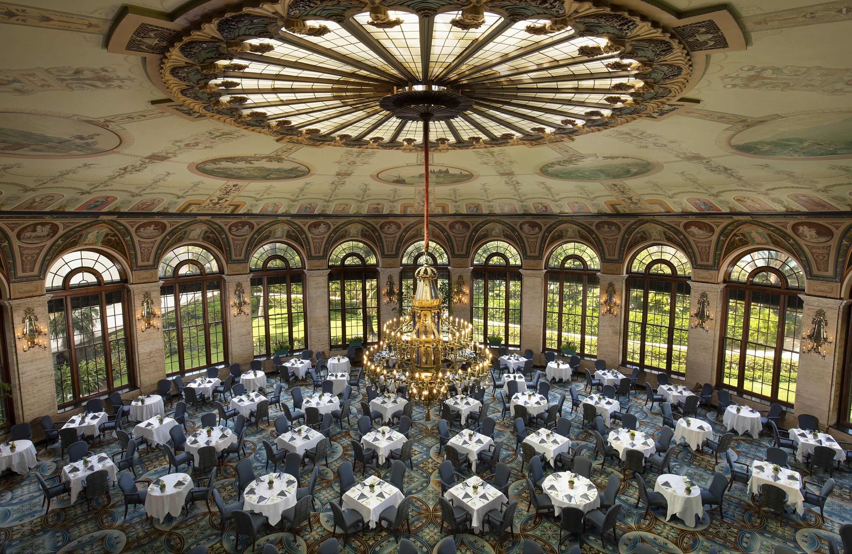A grand hall with a central chandelier, arched windows, and ornate ceiling artwork. Tables are arranged for dining.