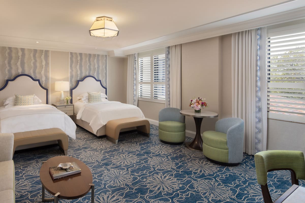 Habitación de hotel con dos camas, una mesita redonda pequeña, una silla y un ventanal grande con persianas.