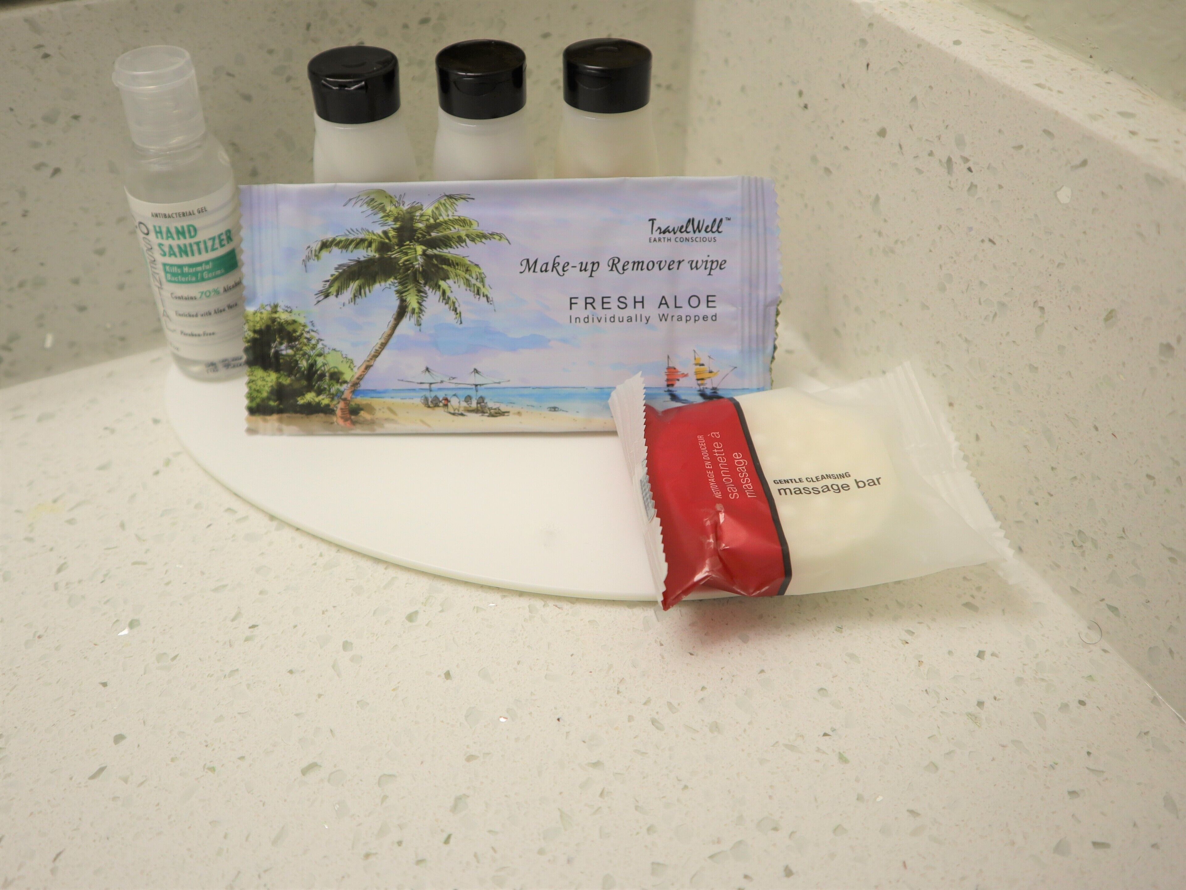 Una encimera de baño con una botella de gel hidroalcohólico, una toallita quitamanchas de maquillaje y una pastilla de jabón roja.