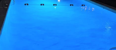 A swimming pool with a blue tint, surrounded by a fence, and a chair visible in the background.