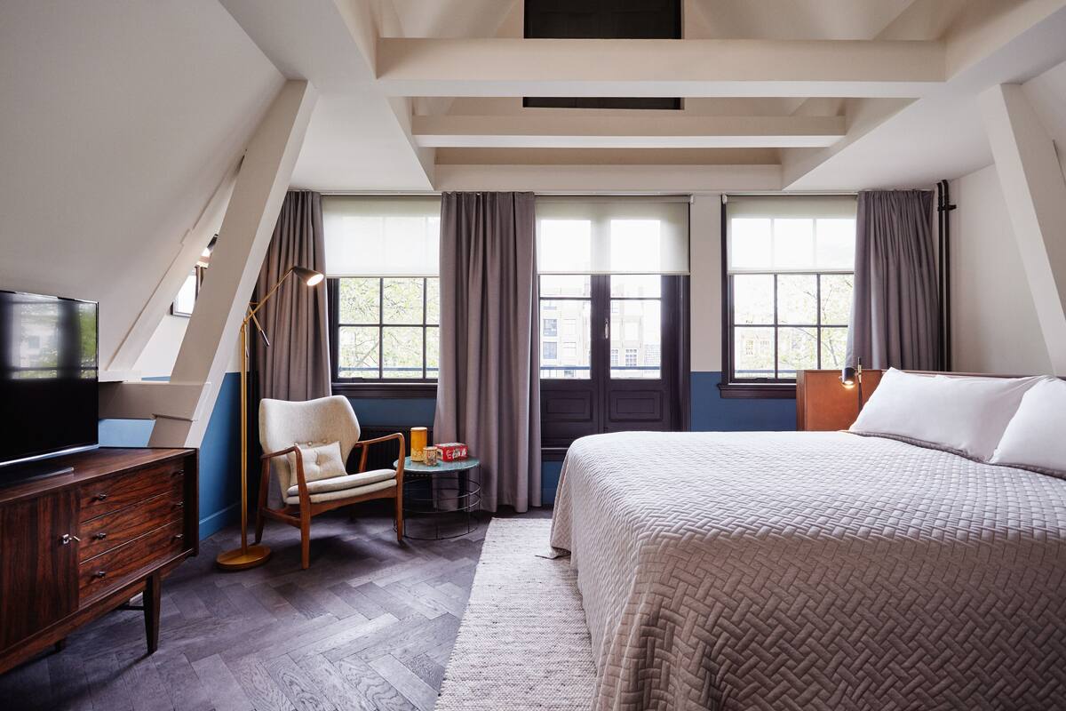 Un dormitorio con una cama grande, televisión, una silla, una mesita y una ventana con cortinas.