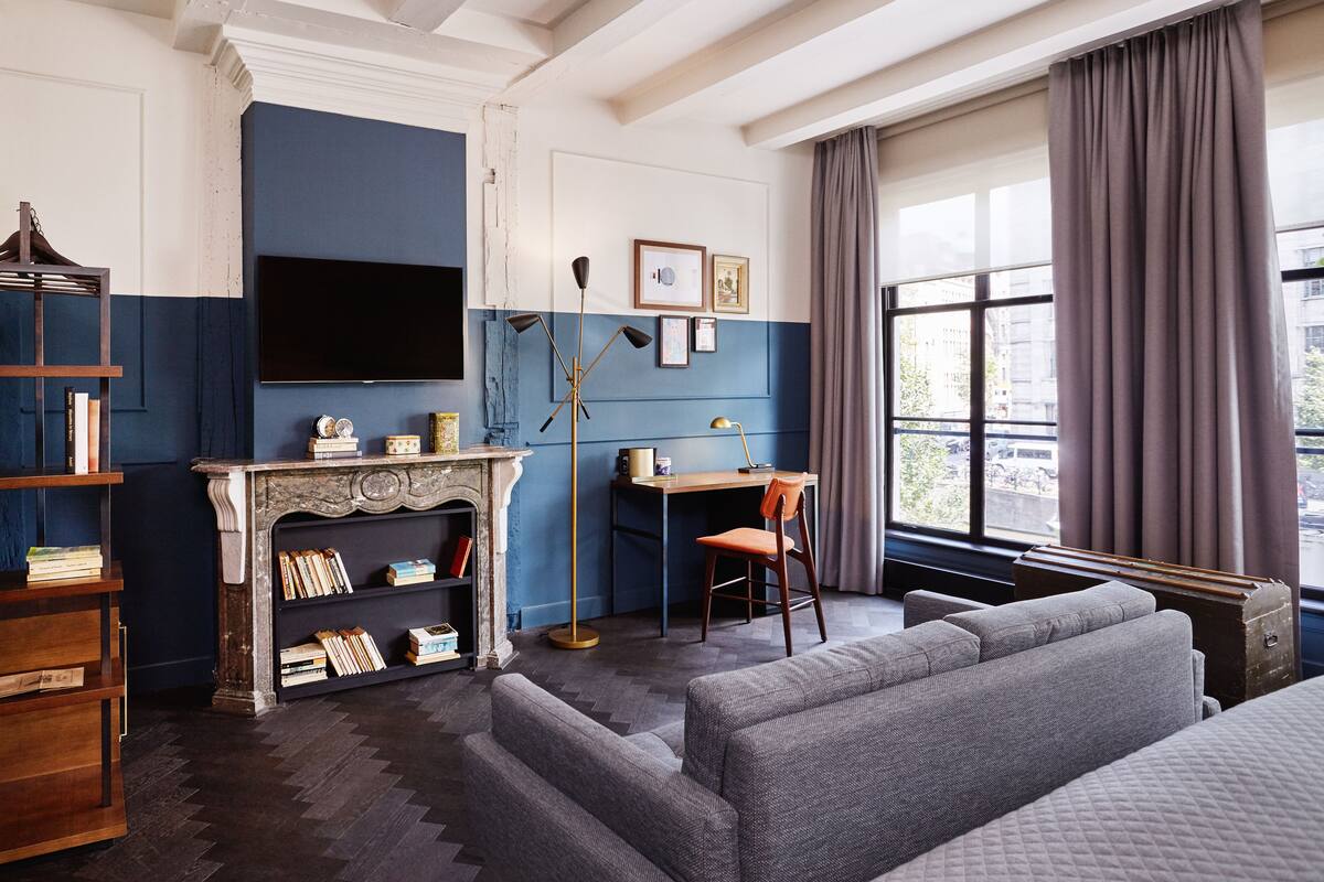 Una habitación con chimenea, un escritorio, una estantería, un sofá y un ventanal con cortinas.