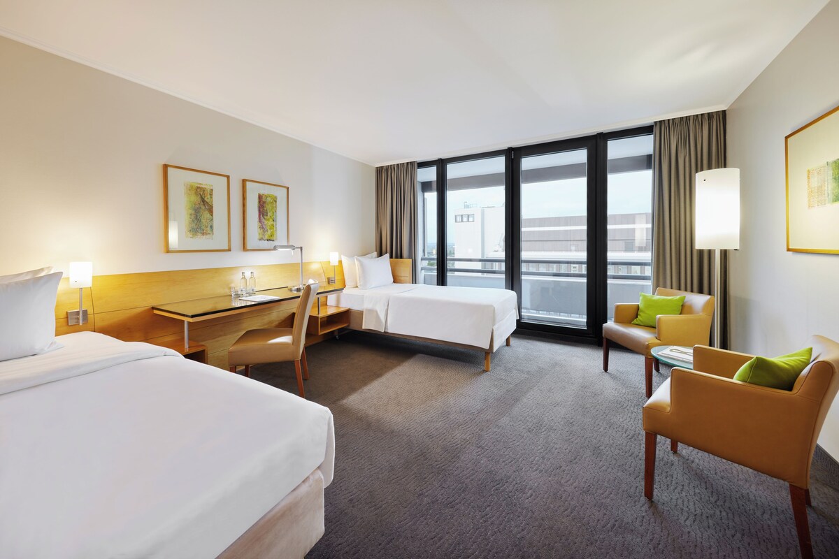 Habitación de hotel con dos camas, un escritorio, una silla y un sofá. Amplias ventanas con cortinas y obras de arte en las paredes.