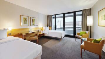 A hotel room with two beds, a desk, a chair, and a sofa. There are large windows with curtains, and artwork on the walls.
