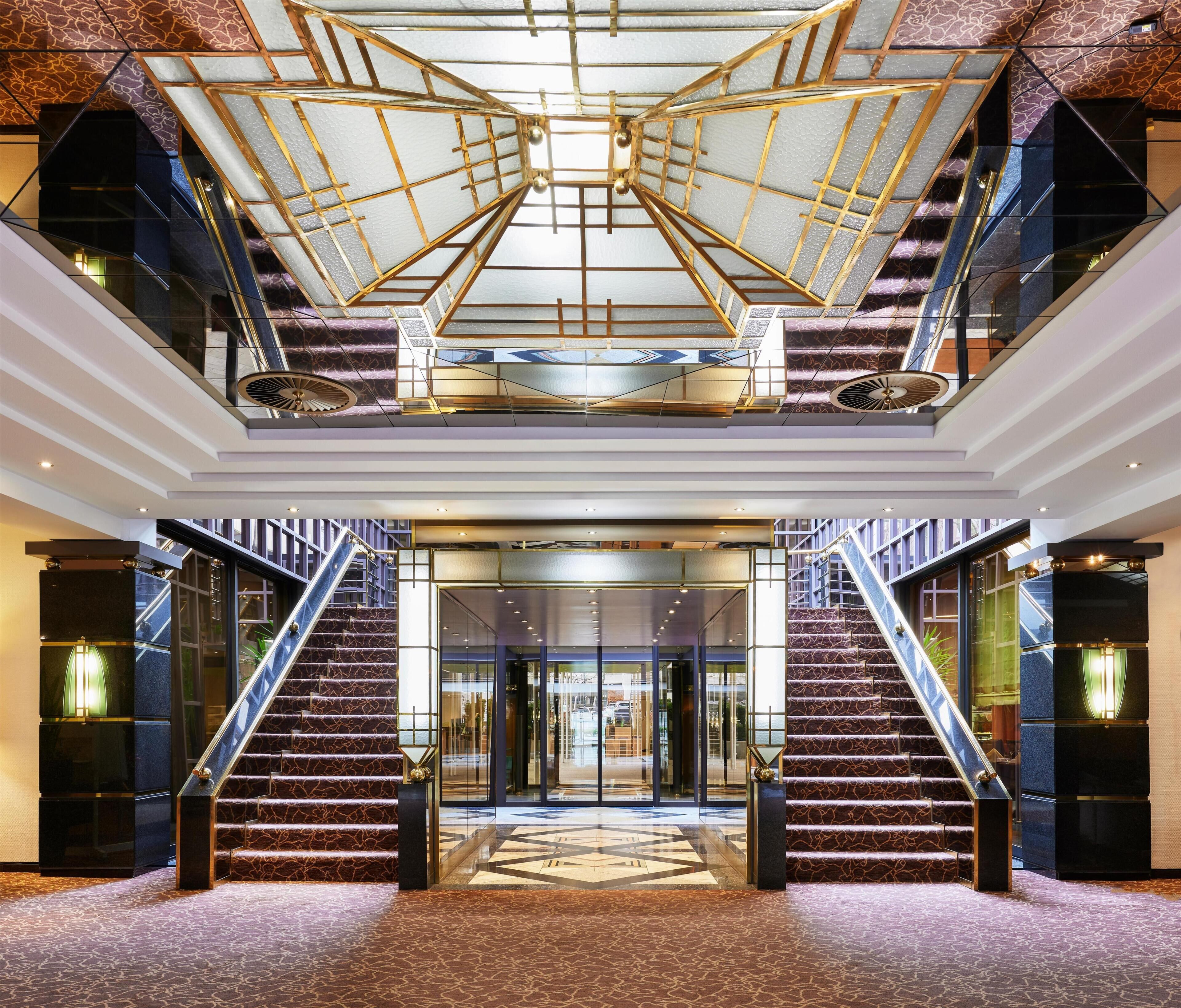 A grand hotel lobby with a central staircase, a glass elevator, and a patterned floor.