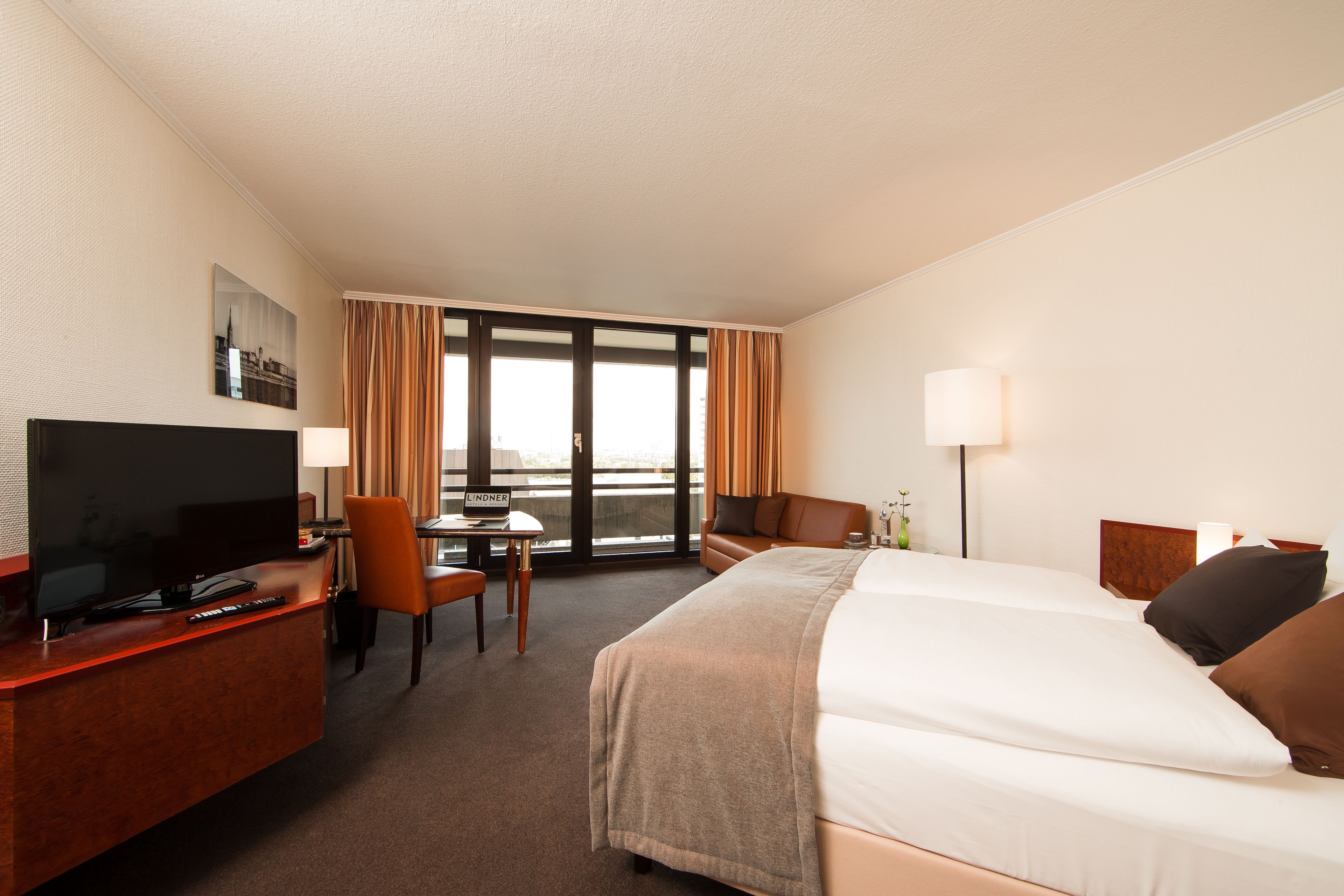 A hotel room with a bed, a desk with a TV, a chair, a table, and a window with curtains.