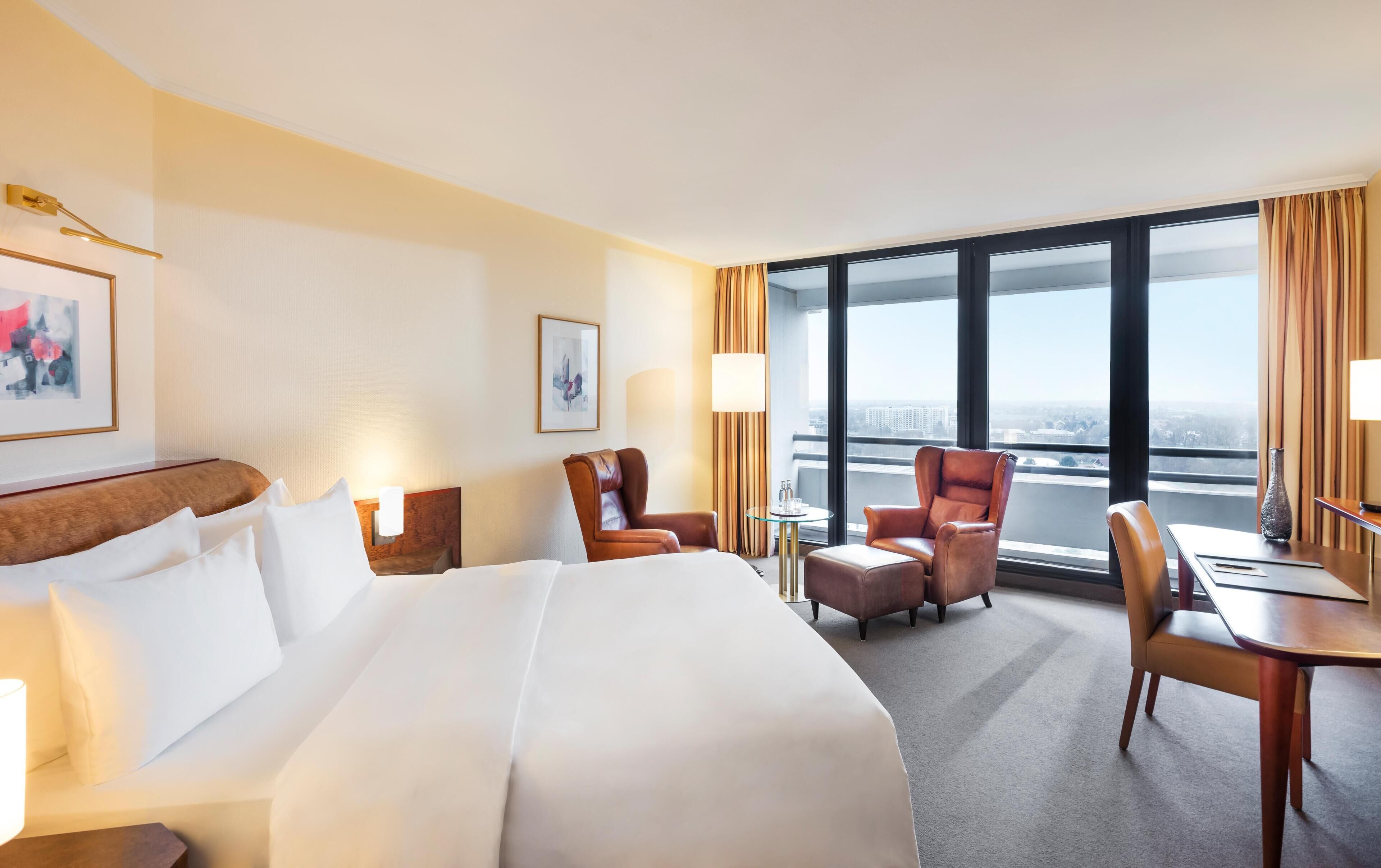 A hotel room with a large bed, two armchairs, a desk, and a view of the city through large windows.
