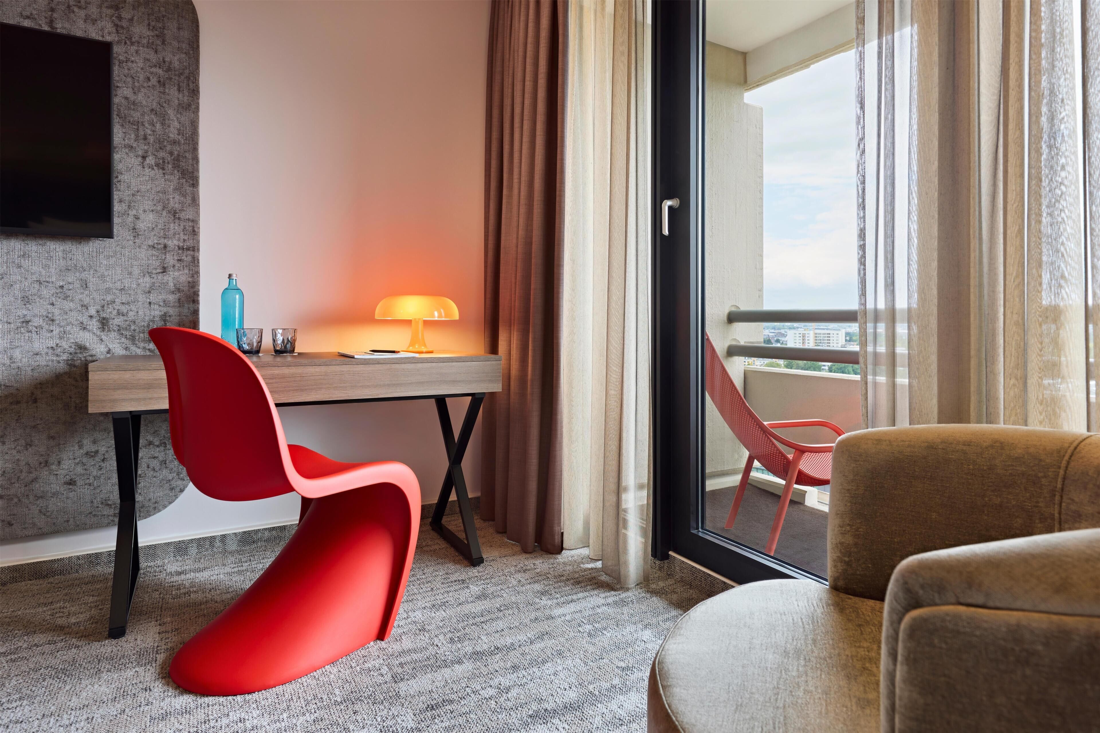 A hotel room with a desk, two chairs, a lamp, and a view of the city through the window.