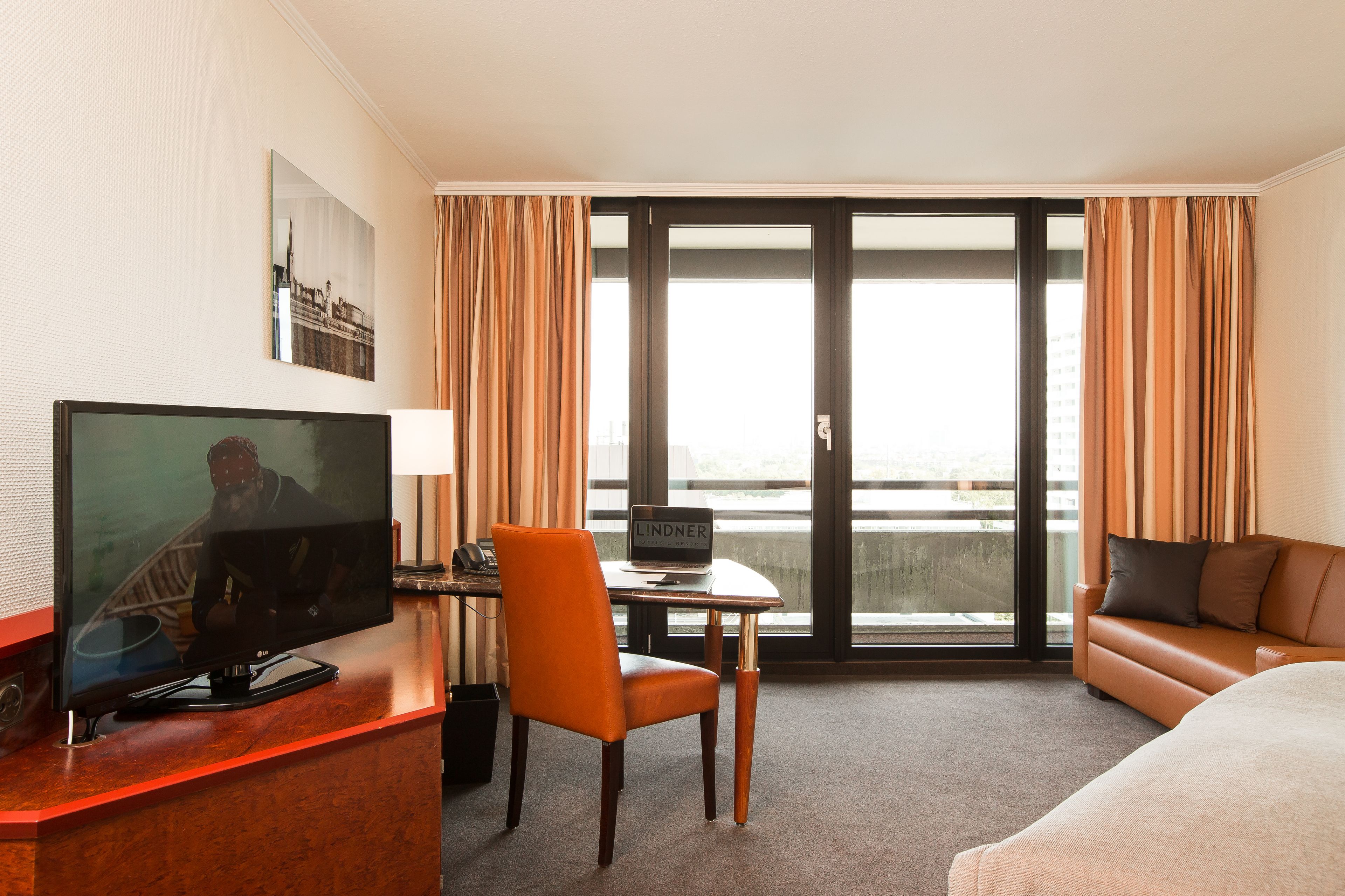 Habitación de hotel con televisión, escritorio, silla, sofá y un amplio ventanal con cortinas.