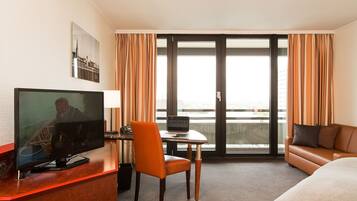 Habitación de hotel con televisión, escritorio, silla, sofá y un amplio ventanal con cortinas.