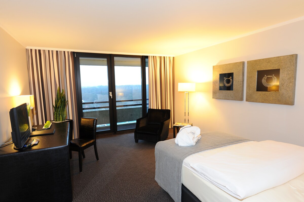 A hotel room with a bed, a desk with a computer, a chair, a lamp, and a view of the outdoors.