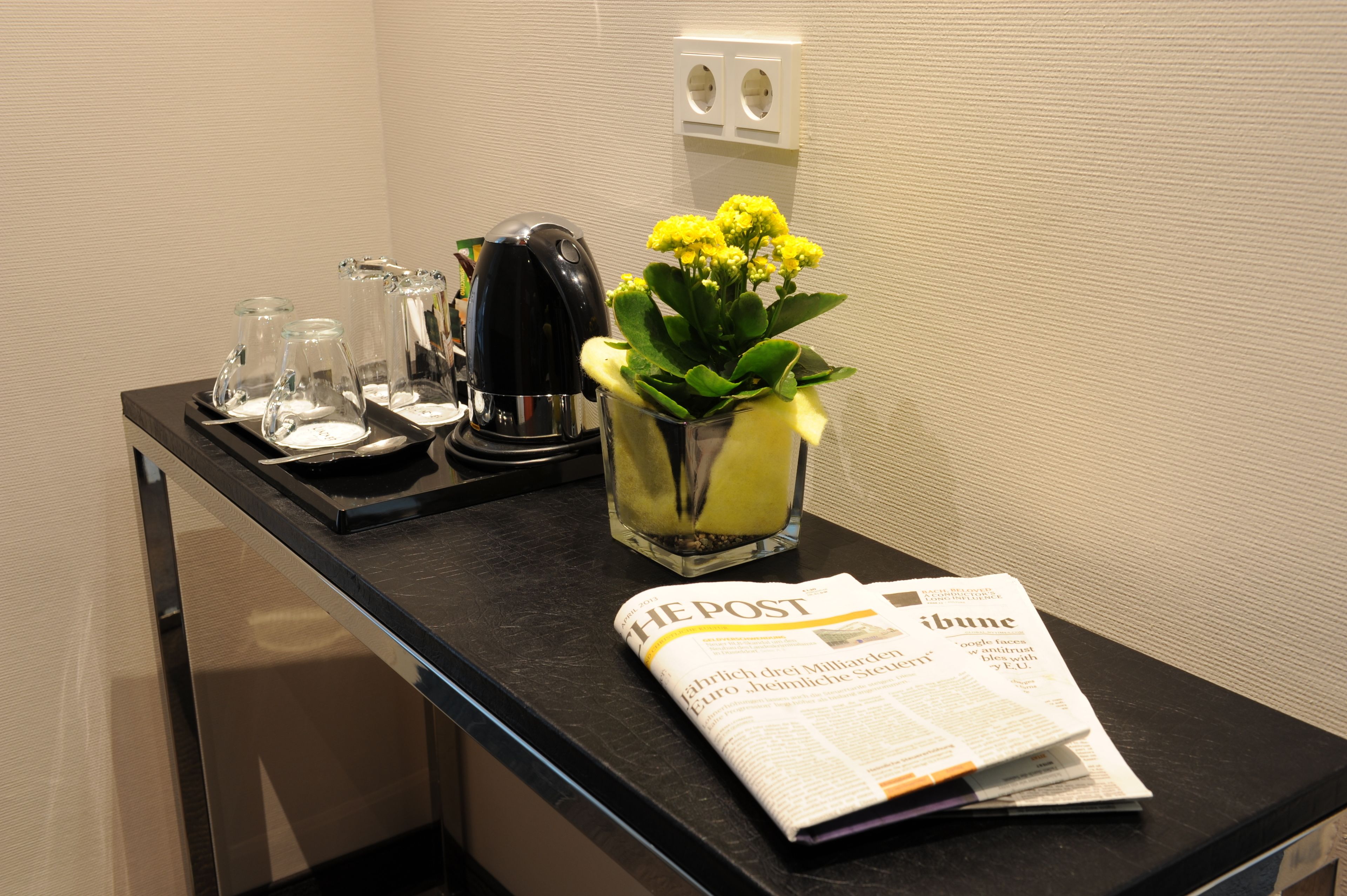 Un comptoir dans une chambre d’hôtel avec un journal, une machine à café et un vase rempli de fleurs jaunes.