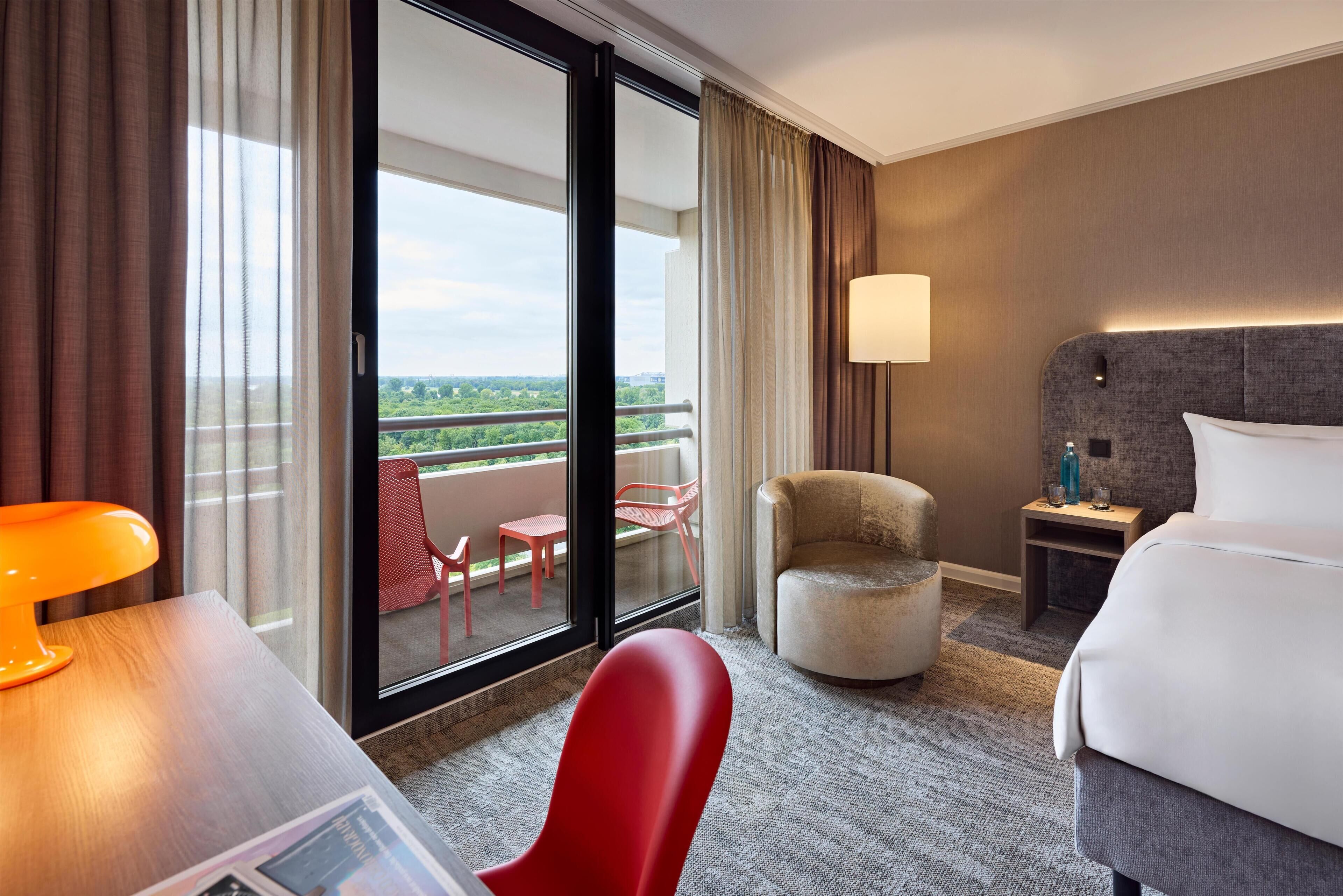 A hotel room with a bed, a desk, a chair, a nightstand, a lamp, and a view of the outdoors.