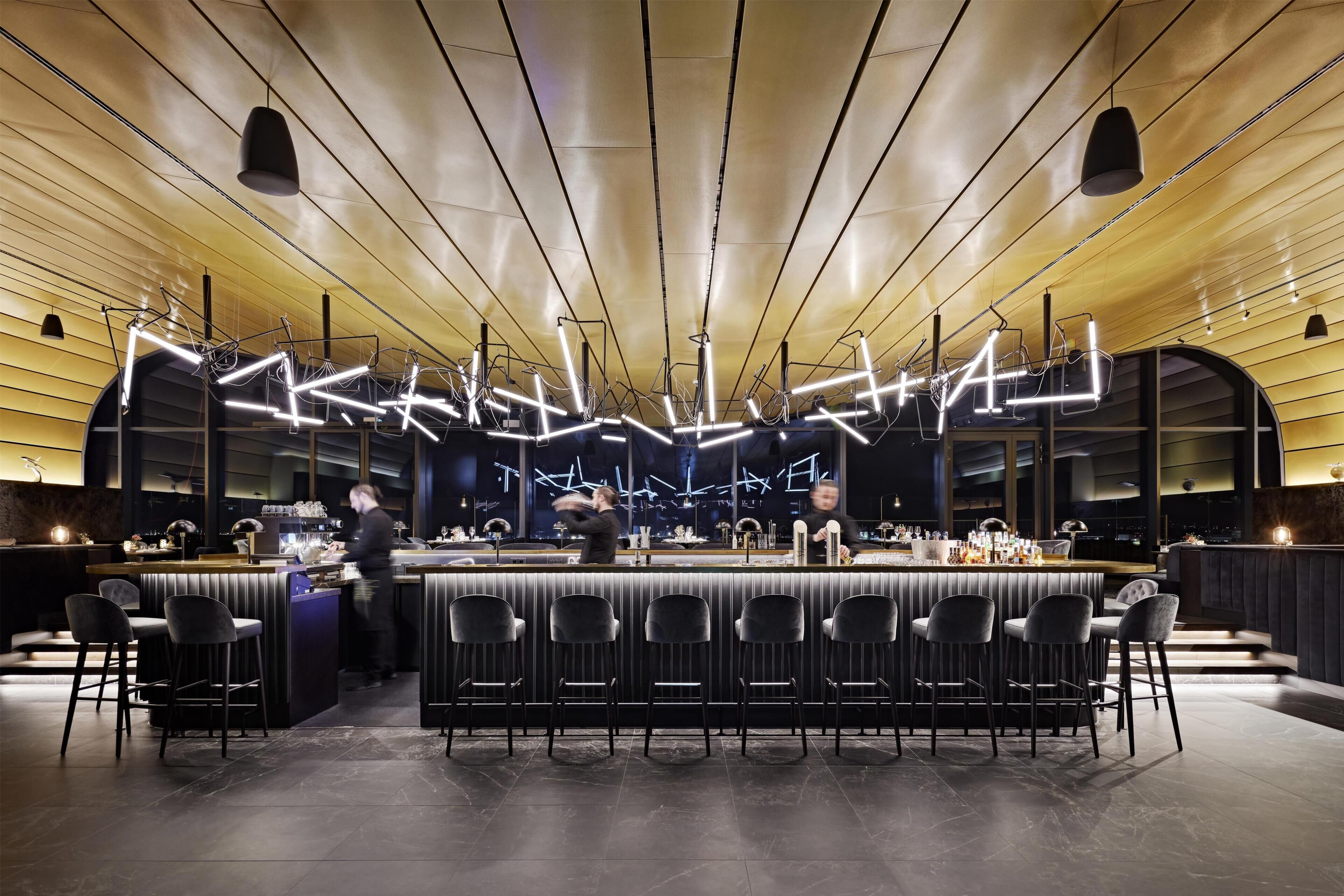 A modern bar with a long counter, multiple bar stools, and a unique ceiling light design.
