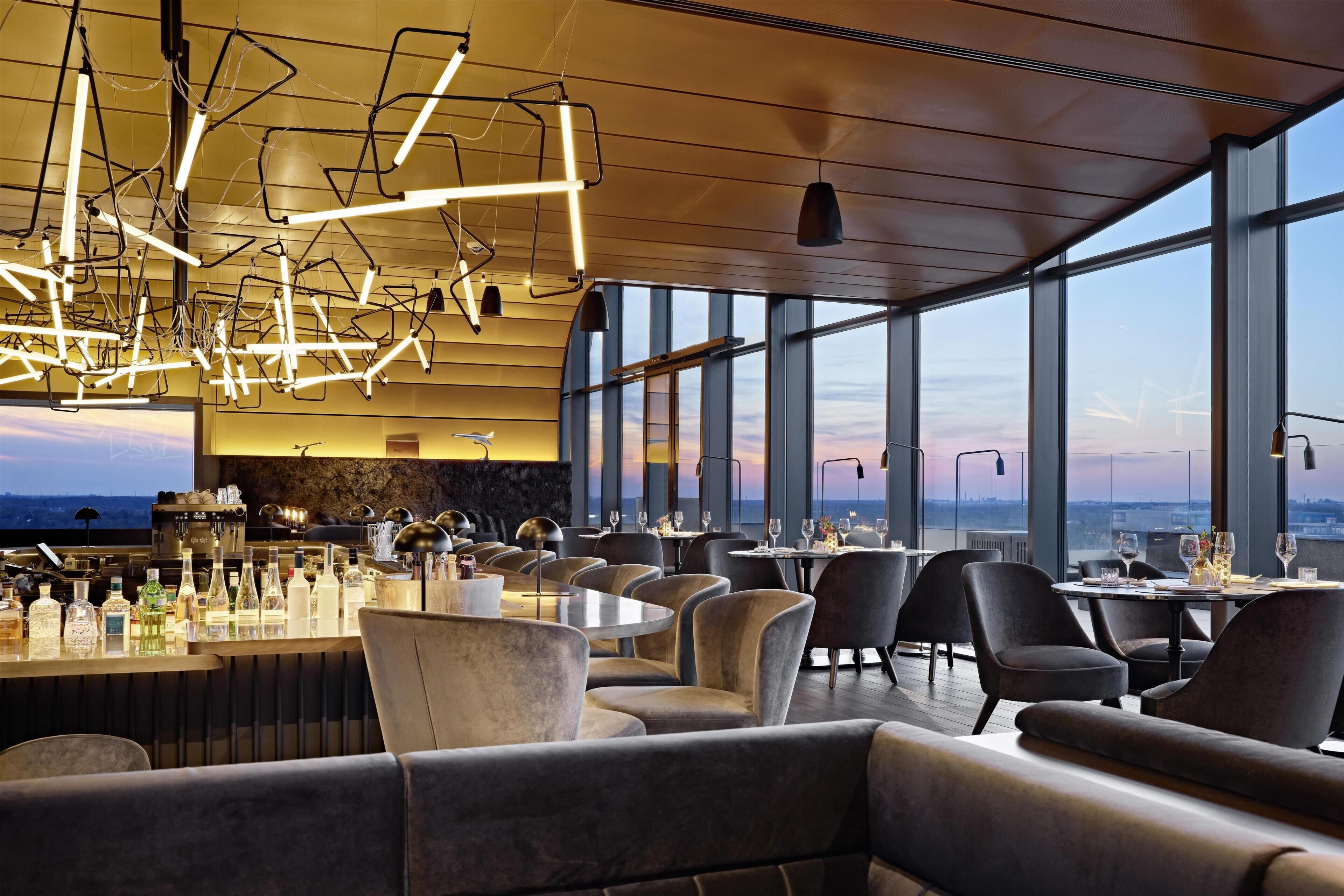 A modern bar area with a high ceiling, pendant lighting, and a view of the cityscape through large windows.