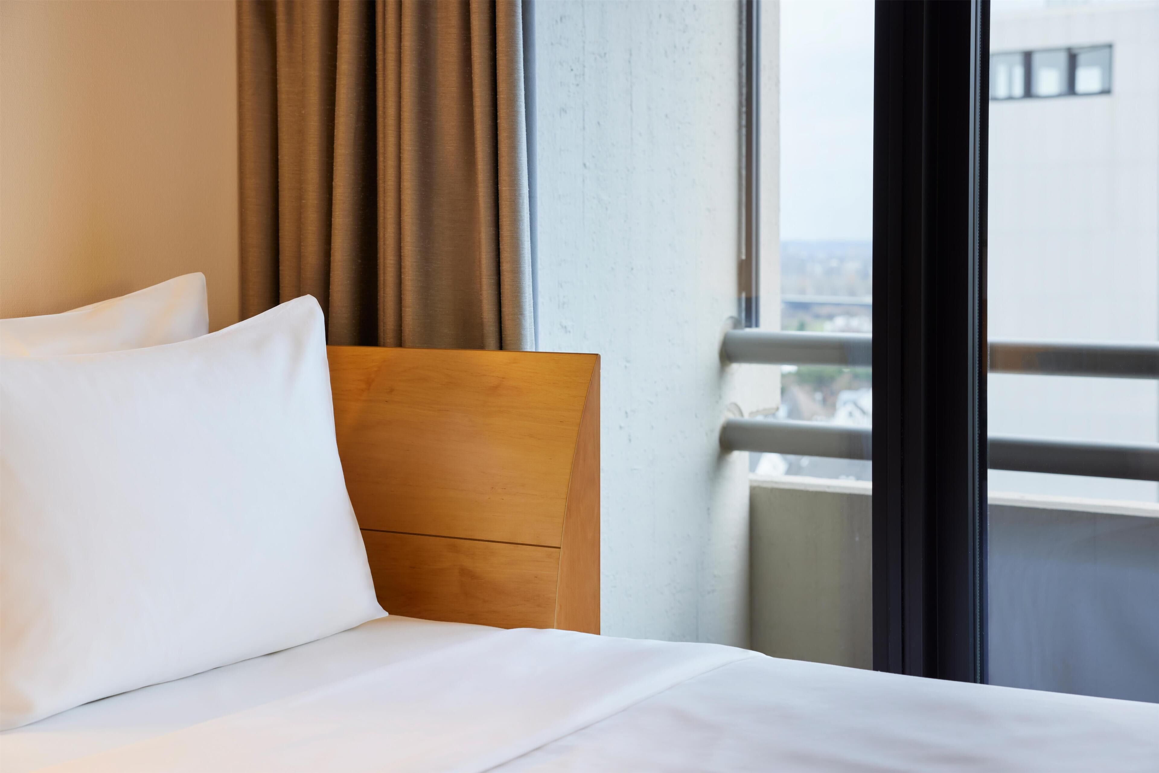 A hotel room with a bed, a wooden headboard, and a view of a cityscape through a window.