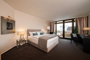 A hotel room with a large bed, a desk, a chair, a lamp, and a view of the city through the window. - Lindner Dusseldorf Seestern, Part of JdV by Hyatt (Düsseldorf)