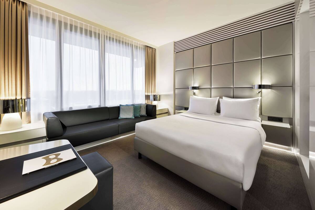 A modern hotel room with a large bed, a sofa, a desk, and a large window with curtains.