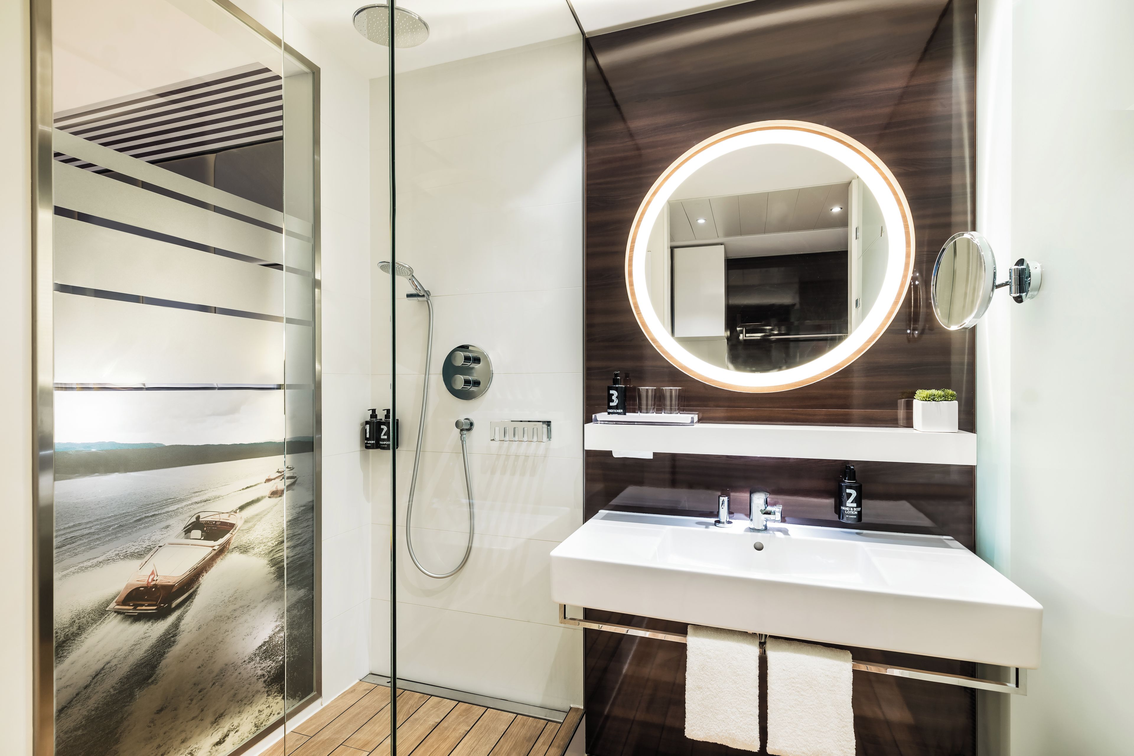 A modern bathroom with a glass shower enclosure, a round mirror with a light, and a white sink.