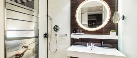 A modern bathroom with a glass shower enclosure, a round mirror with a light, and a white sink.