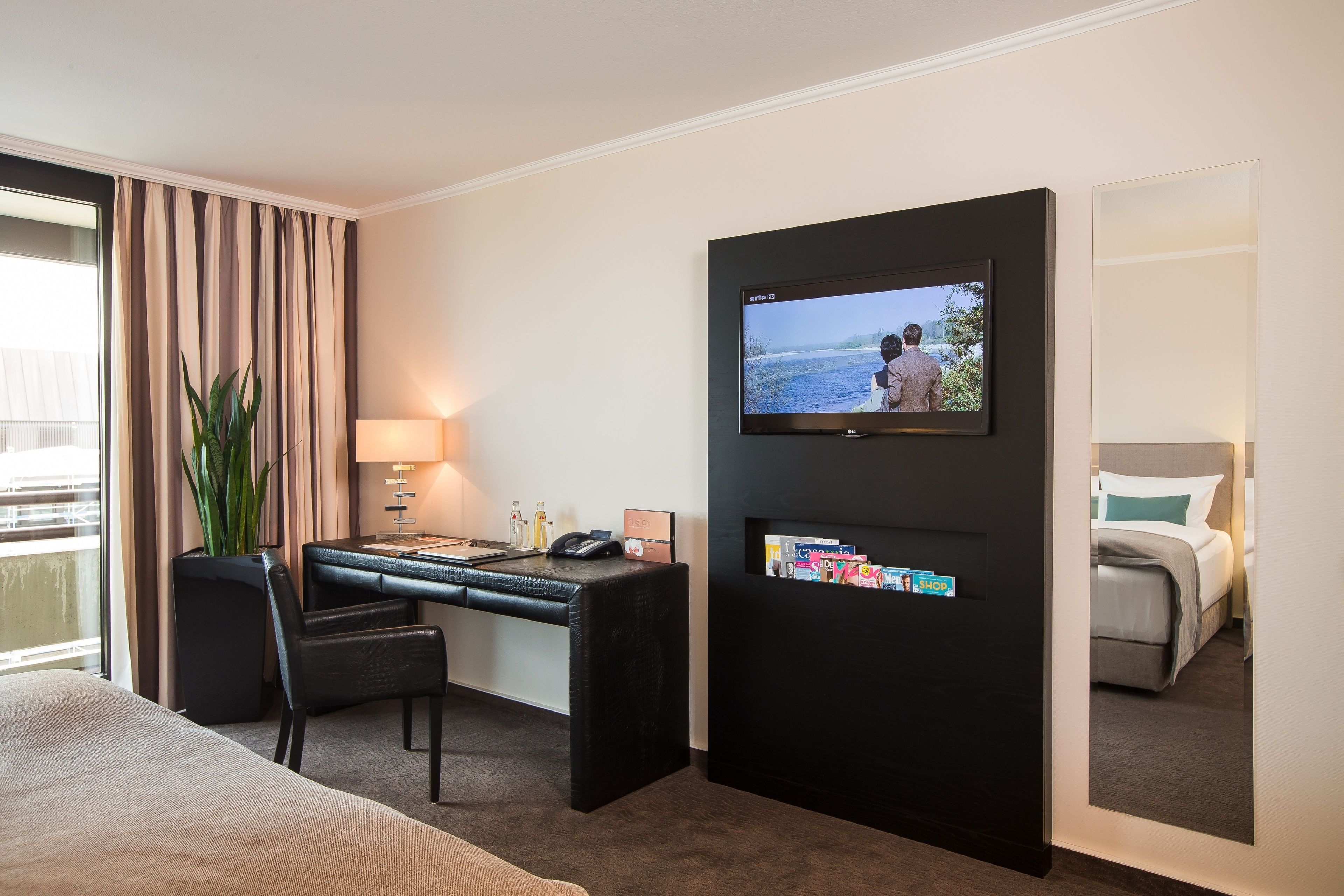 Habitación de hotel con cama, escritorio con silla, televisor montado en la pared y un amplio ventanal con cortinas.