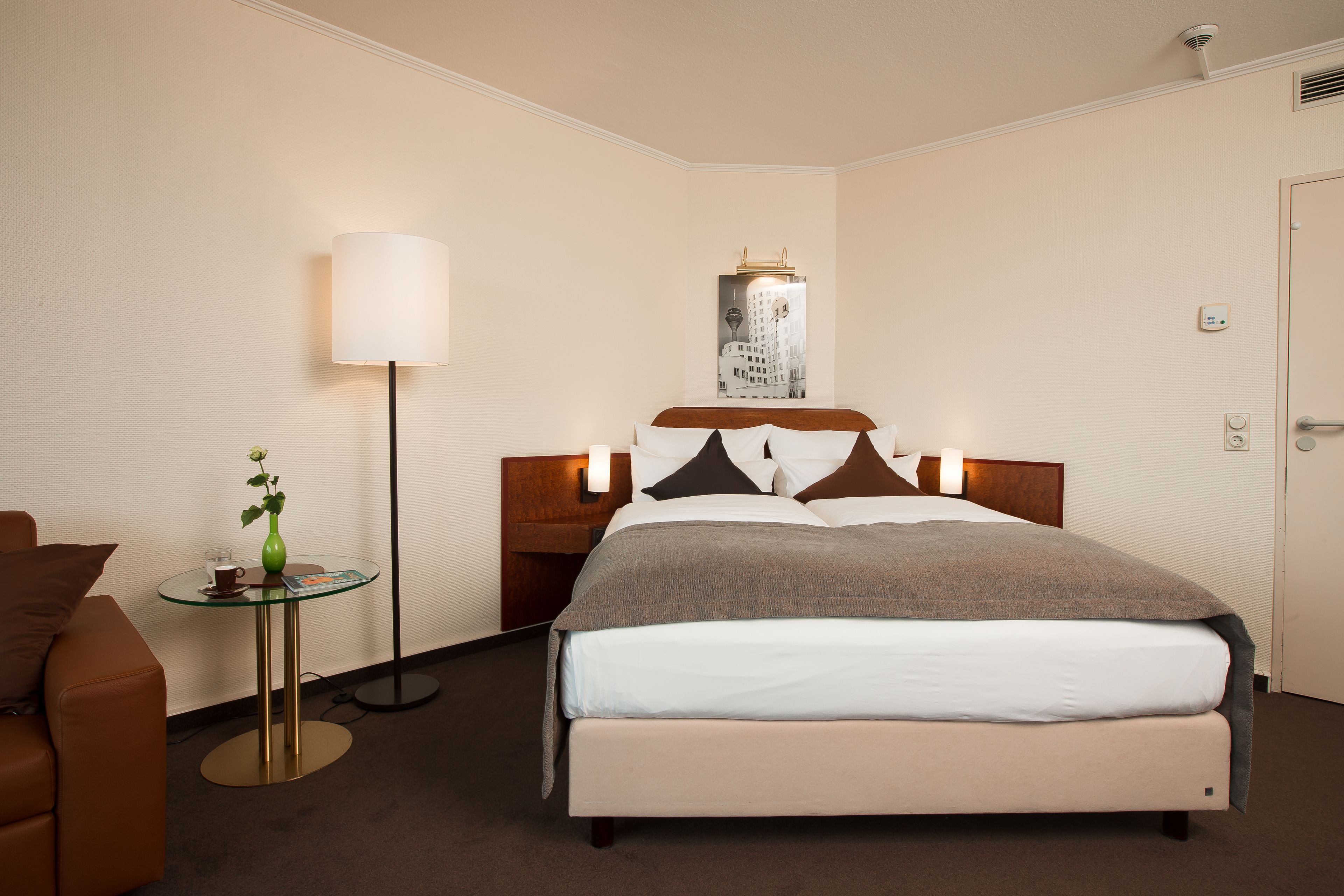 A hotel room with a bed, a bedside table with a lamp, a small table with a vase, and a chair.