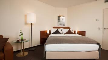 A hotel room with a bed, a bedside table with a lamp, a small table with a vase, and a chair.